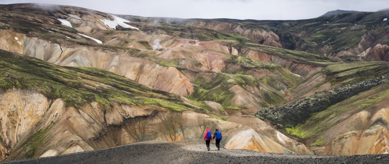 Two persons walking on a trail surrounded by orange and green hills.